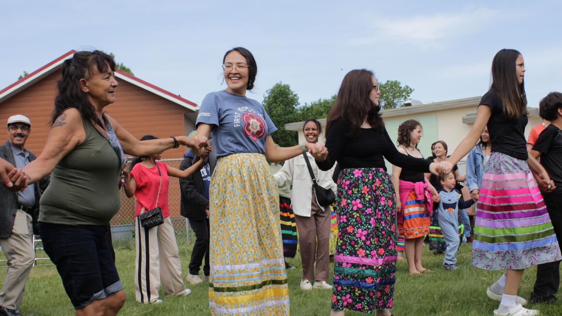 The image shows a group of people gathered outside on a lawn, participating in what appears to be a group dance or community activity. The participants are arranged in a circle or line, holding hands. Several women are wearing long, colourful skirts with floral or geometric patterns and bright colours (yellow, blue, pink, green) reminiscent of traditional dress. Adults, as well as a few children, are participating or observing the scene. Some people are wearing more modern outfits.