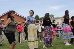 The image shows a group of people gathered outside on a lawn, participating in what appears to be a group dance or community activity. The participants are arranged in a circle or line, holding hands. Several women are wearing long, colourful skirts with floral or geometric patterns and bright colours (yellow, blue, pink, green) reminiscent of traditional dress. Adults, as well as a few children, are participating or observing the scene. Some people are wearing more modern outfits.
